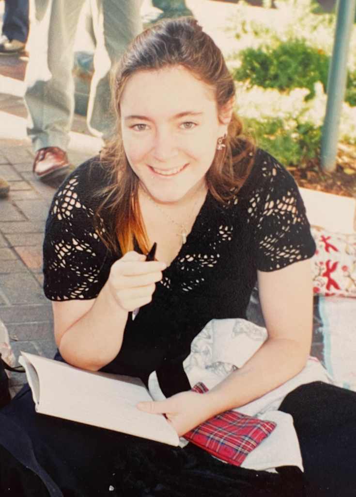 Smiling woman sitting with a book on her lap, pointing a pen at the camera