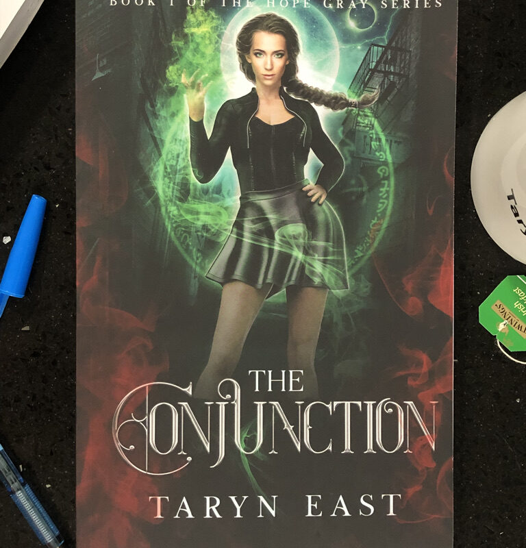 Paperback book (The Conjunction by Taryn East) on black table. The edge of a pen, a tea-cup and a book with hand-written notes are visible around the edges.