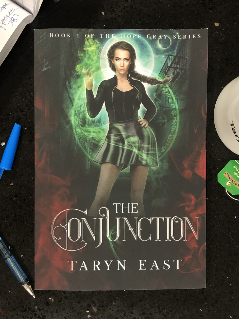 Paperback book (The Conjunction by Taryn East) on black table. The edge of a pen, a tea-cup and a book with hand-written notes are visible around the edges.